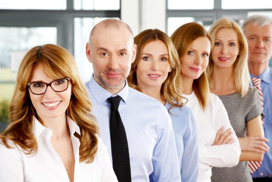 Image of business people standing at office in a row. Portrait of mixed age businesswomen and businessmen standing with arms crossed and looking at camera.